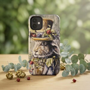 May include: A smartphone case featuring a detailed watercolor illustration of a rabbit wearing a top hat adorned with berries and foliage. The artwork displays muted earth tones, including browns, greens, and deep reds, with a visible artist signature.
