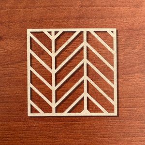 May include: Square, off-white coaster with a geometric chevron pattern. The coaster is made of a light-coloured material and sits on a dark brown wooden surface. The design features a series of diagonal lines forming a repeating V-shape.