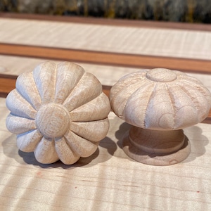 May include: Two unfinished wooden cabinet knobs. One knob is shaped like a flower with detailed petal carvings. The other knob has a mushroom-like design with a rounded top and a pedestal base. Both knobs are made of light-colored wood.