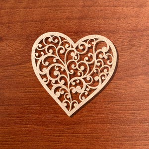 May include: A laser-cut wooden heart with an intricate, swirling filigree design. The heart is a light beige color and sits on a dark brown wooden surface. The detailed pattern creates a delicate, openwork effect, perfect for crafts or decor.