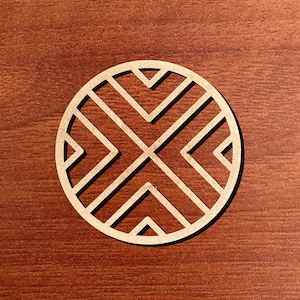 May include: A round, light-colored wooden coaster with a geometric design. The coaster features a series of intersecting lines and angles, creating a symmetrical pattern. The coaster is set against a dark wood grain background.