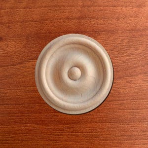May include: A light-colored, unfinished wooden rosette with a circular design. The rosette features concentric rings and a central, raised button. The wood grain is visible, and the rosette is set against a reddish-brown background.