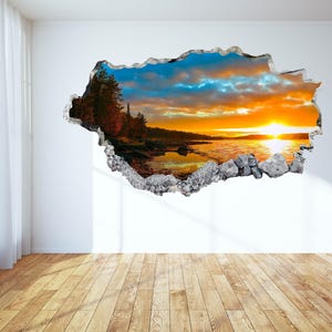 May include: A wall decal featuring a vibrant sunset over a lake, framed by a torn wall effect. The scene includes a bright orange sun, a reflective lake, and a forested shoreline. The decal is set against a white wall with a wooden floor.