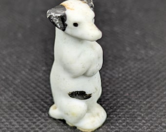 Puede incluir: Una pequeña figurita de perro de porcelana blanca con marcas negras. El perro está de pie sobre sus cuatro patas y tiene la cabeza inclinada hacia un lado.
