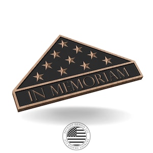 Könnte beinhalten: Eine bronzefarbene Gedenktafel in Form einer gefalteten amerikanischen Flagge mit 13 Sternen und der Inschrift "IN MEMORIAM" in erhabenen Buchstaben. Die Tafel hat einen dunklen Hintergrund und einen hellbraunen Rand. Unten befindet sich ein rundes Logo mit der amerikanischen Flagge.
