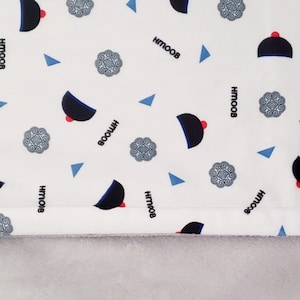 May include: A white fabric baby blanket with a repeating pattern of blue and black hats, blue triangles, and gray floral designs. The blanket has a white border and a gray fleece backing with the text "Kab Yeeb Xyooj" embroidered in black.