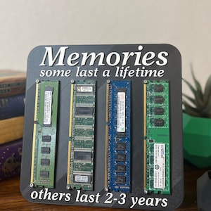 May include: A black sign with the text "Memories some last a lifetime others last 2-3 years". Four computer memory sticks are mounted on the sign. The memory sticks are green, blue, and black.