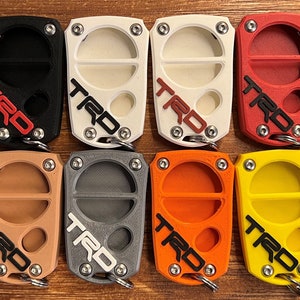 May include: A selection of keychains in a range of colours: black, white, red, green, tan, grey, orange, yellow, and blue. Each keychain has the letters "TRD" in a contrasting colour and a metal keyring.