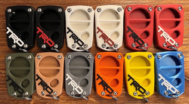 May include: A selection of keychains in a range of colours: black, white, red, green, tan, grey, orange, yellow, and blue. Each keychain has the letters "TRD" in a contrasting colour and a metal keyring.