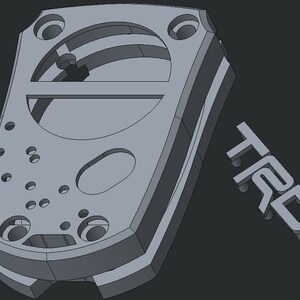 May include: A grey, 3D-rendered component with multiple holes and a curved design. The letters "TRD" are displayed separately, suggesting a mechanical or automotive part. The overall design is geometric and industrial.