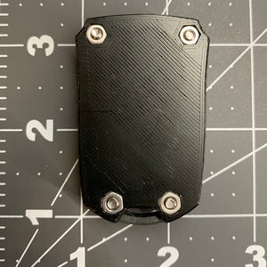 May include: A black, rectangular object with a textured surface and four silver bolts. The object has a small, curved blade at the bottom. The background is a grey cutting mat with white grid lines and numbers.