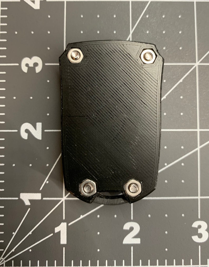 May include: A black, rectangular object with a textured surface and four silver bolts. The object has a small, curved blade at the bottom. The background is a grey cutting mat with white grid lines and numbers.