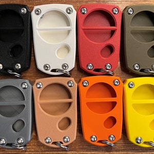May include: Eight key fobs in a range of colours: black, white, red, olive green, grey, tan, orange, and yellow. Each fob has a rectangular form with a circular opening and a metal keyring attachment. The fobs are secured with four small screws.