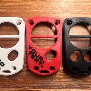 May include: Three 3D-printed keychains in white, red, and black. The white keychain has the text "MY TACO", the red one says "YOUR TEXT", and the black one is blank. Each has a circular design with holes.
