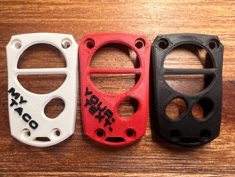 May include: Three 3D-printed keychains in white, red, and black. The white keychain has the text "MY TACO", the red one says "YOUR TEXT", and the black one is blank. Each has a circular design with holes.