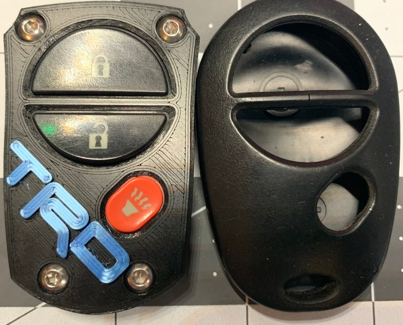 May include: Two black car key fobs. One has a blue "TRD" logo and an orange button. The other is a plain black fob. Both have buttons for locking and unlocking the car.