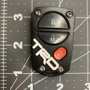 May include: Black car key fob with the white letters "TRD" and a red button. The fob has two black buttons with lock and unlock symbols. The fob is secured with four silver screws.
