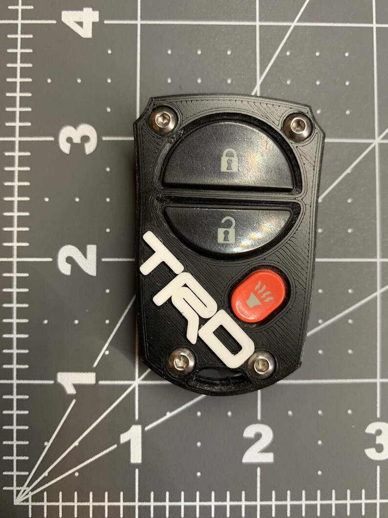 May include: Black car key fob with the white letters "TRD" and a red button. The fob has two black buttons with lock and unlock symbols. The fob is secured with four silver screws.