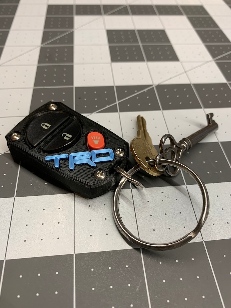 May include: Black key fob with blue "TRD" lettering and a red button. The fob is attached to a silver key ring with two keys and an antique key. The key fob has two black buttons with lock and unlock symbols.