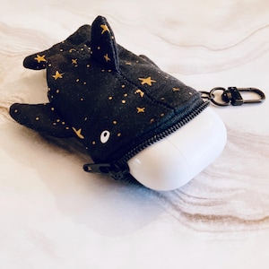 May include: A black fabric AirPod case shaped like a whale, with gold star accents. The case has a zipper closure and a small metal clasp. The white AirPods are partially visible.
