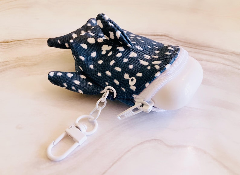 May include: A whimsical, navy blue and white polka-dotted whale shark-shaped pouch with a white zip and a white metal clasp. The pouch has small fins and a rounded white base. The pouch is designed to hold small items.