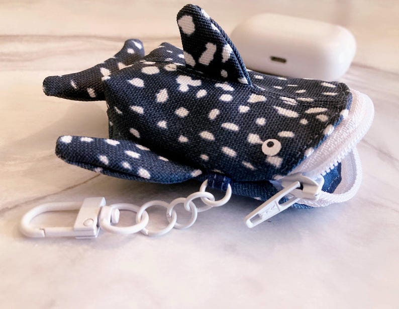 May include: A navy blue and white polka-dotted whale shark-shaped case with a white zip and a white metal clasp. The case is designed to hold small items, such as earbuds. A white earbud case is in the background.