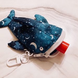 May include: A blue whale-shaped pouch with a celestial pattern of white stars and crescent moons. The pouch has a white zipper and a white clip. A red tube is partially visible. The pouch is designed to hold small items.