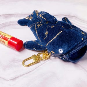 May include: A navy blue whale-shaped coin purse with gold accents and a gold-colored keychain. Next to it is a tube of Burt's Bees Strawberry moisturizing lip balm with a red cap.