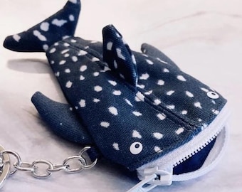 May include: A whale shark shaped coin purse made of dark blue fabric with white polka dot patterns. The pouch features a white zipper closure along the mouth, a dorsal fin, side fins, and a tail. A silver metal chain and key ring are attached.
