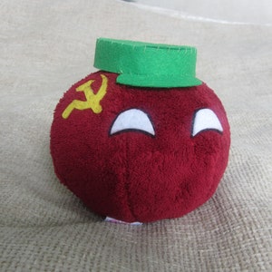 Op de afbeelding: Rood pluche speelgoed in de vorm van een bol met een geel hamer-en-sikkel symbool aan de zijkant. Het speelgoed heeft een wit gezicht met twee witte ogen en een groene hoed op de top.