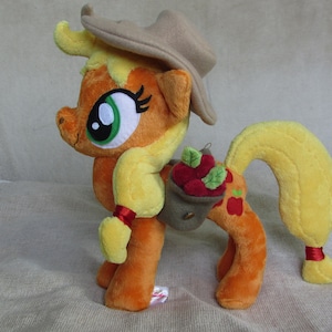Puede incluir: Un juguete de peluche de Applejack de My Little Pony: La Magia de la Amistad. El juguete es naranja y amarillo con un sombrero marrón y una cesta de manzanas en la espalda.