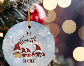 May include: A round Christmas ornament with a gold string, featuring a family of gnomes wearing red hats. The ornament has the text "The Smith Family 2025" in a decorative font, with snowflakes in the background. The ornament is hanging on a Christmas tree.