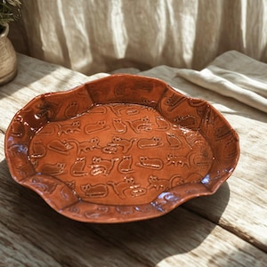 May include: A terracotta-colored ceramic serving platter with a wavy edge. The platter is decorated with an embossed pattern of stylized cats in various poses. The glossy finish enhances the warm, earthy tones of the dish.