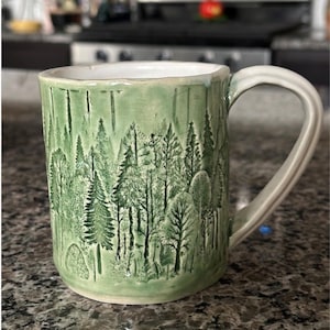May include: A ceramic mug with a light green glaze featuring a detailed, embossed forest scene with various evergreen and deciduous trees. The mug has a white interior, a sturdy handle, and a textured, hand-crafted appearance with visible relief.