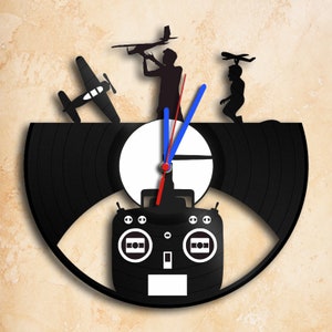 Può includere: Un orologio in vinile nero con quadrante bianco e lancette nere. L'orologio è decorato con silhouette di persone che fanno volare aerei radiocomandati e un telecomando.