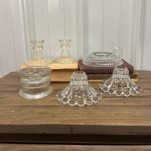 May include: Five clear glass candle holders of various styles and sizes. The candle holders are arranged on a wooden surface.