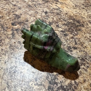 May include: A carved dragon head figurine in shades of green and black. The sculpture is made of a stone material with a polished finish. The dragon's head is detailed with scales and a defined snout.