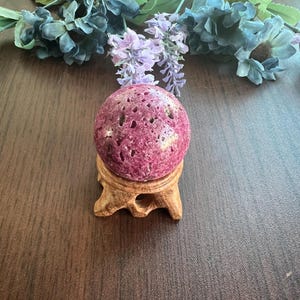 May include: A deep pink, porous sphere rests on a small, carved wooden stand. The sphere is the focal point, with a textured surface and a rich, reddish-purple hue. Decorative floral accents in shades of blue, green, and purple are arranged in the background.