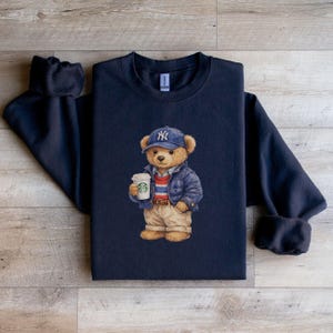 May include: Navy blue sweatshirt featuring a cartoon bear wearing a blue baseball cap with the NY logo, a blue jacket, and khaki pants. The bear is holding a coffee cup. The sweatshirt has a crew neck and long sleeves.