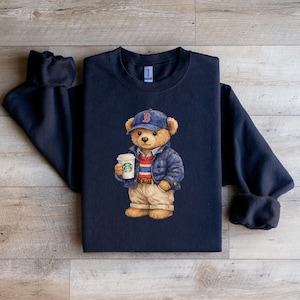 May include: Navy blue sweatshirt with a watercolour illustration of a teddy bear wearing a blue baseball cap with a red "B", a blue jacket, and khaki trousers. The bear holds a coffee cup with a green logo. The sweatshirt has a crew neck.