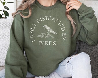 May include: A green sweatshirt with the text "Easily Distracted By Birds" in white lettering. A small bird is perched on a branch with leaves.