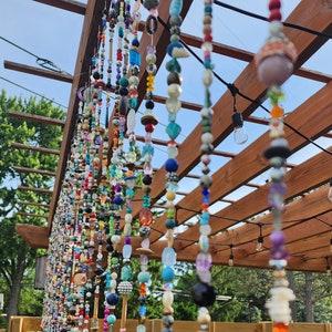 May include: A colorful beaded curtain hangs from a wooden pergola. The beads are strung on multiple strands and are made of various materials, including glass, wood, and metal. The curtain is a vibrant mix of colors, including red, blue, green, yellow, and purple.
