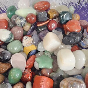 May include: A collection of polished gemstones in various shapes and colors, including red, green, white, and brown. Some stones are heart-shaped or star-shaped. The image is taken from above, showing the stones filling the frame.