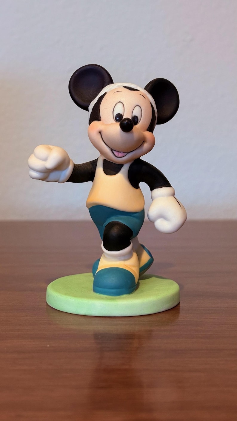 May include: A figurine of Mickey Mouse in a running pose. Mickey is wearing a yellow shirt, blue shorts, and blue and yellow shoes. He has white gloves and a white headband. The figurine is on a green base.