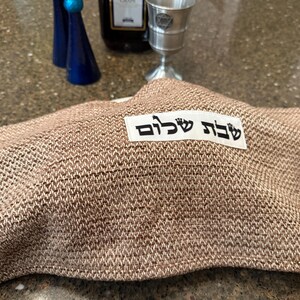May include: A woven brown challah cover with the Hebrew text "Shabbat Shalom" on a white label. The cover is draped over a loaf of bread. In the background are blue decorative bottles, a wine bottle, and a silver cup.