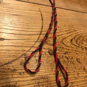 May include: Two strands of twisted cord, one red and black, the other black and red, lie on a wooden surface. Each cord is looped at the end, creating a teardrop shape. The cords are intertwined.
