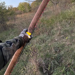 May include: A wooden bow with a pink string and a yellow arrow rest. The bow is held by a person wearing a camouflage sleeve.