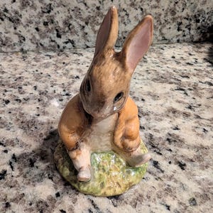 May include: A ceramic figurine of a brown rabbit with long ears, wearing a tan jacket, sitting on a textured green base. The rabbit has dark eyes, a light-colored chest, and a glossy finish, depicted in a seated position with its legs extended forward.