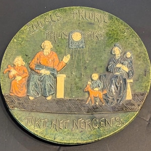 May include: A green ceramic plate with a raised design of a family sitting around a clock. The plate has the text "Zoo als Tklokje Thuis Tikt Tikt Het Nergens" around the edge.