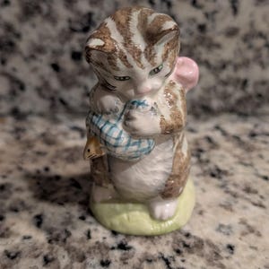 May include: A ceramic figurine of a brown and white tabby kitten standing on a pale green base. The kitten has green eyes and is holding a blue and white checkered bundle in its front paws, with a small pink bow visible behind its head.